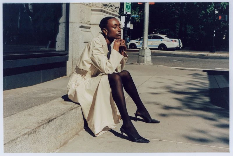 A woman sits on steps outdoors wearing a tan trench coat, black tights, and heels, with a blurred police car in the background.