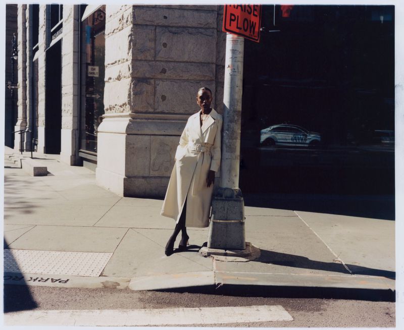 An African-American woman in a light coat poses against a pillar in a city. Modern fashion meets urban architecture in natural lighting.