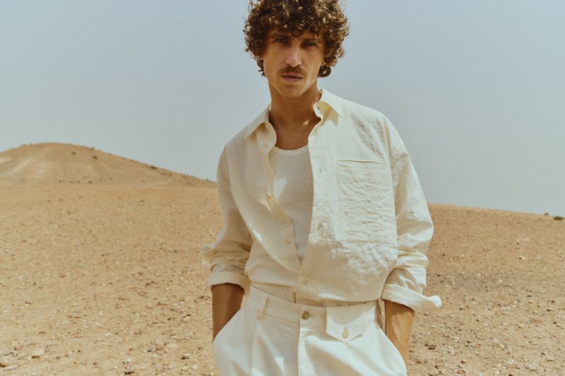 Man in an open linen shirt poses in a desert landscape with a small sand dune hill and pale sky as a warm, natural light source.