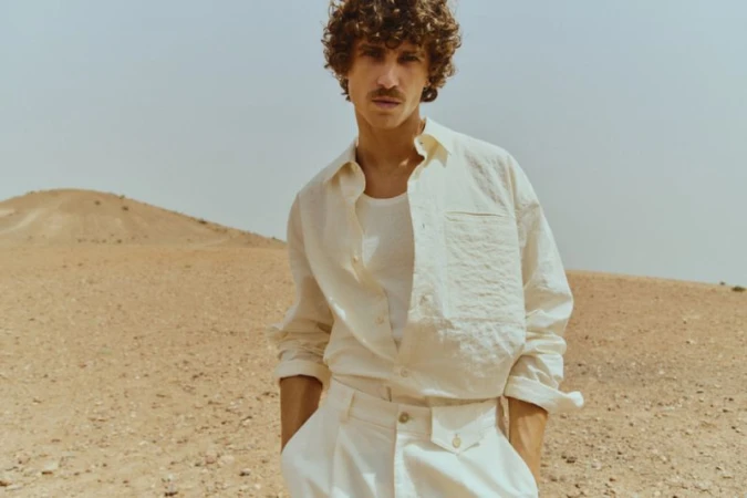 Man in an open linen shirt poses in a desert landscape with a small sand dune hill and pale sky as a warm, natural light source.