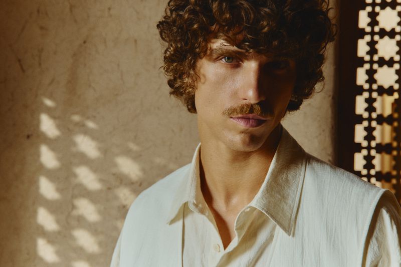 Portrait of a young adult male with curly brown hair and a mustache, wearing a light collared shirt, against a patterned tan background.