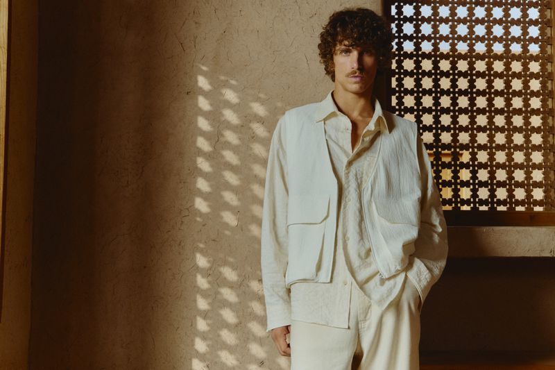 A man poses in an off-white linen outfit in front of a stucco wall with sunlight shining through a lattice window, creating shadows.