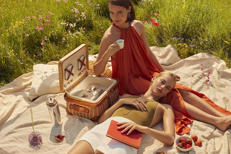 Two women enjoy a picnic outdoors on a blanket in a sunny field with a picnic basket, book, and refreshments, creating a lifestyle scene.