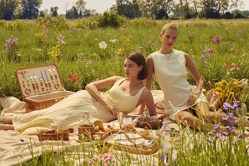 Two adult women enjoy a sunlit picnic in a wildflower meadow. They are seated on a blanket with a picnic basket and food.