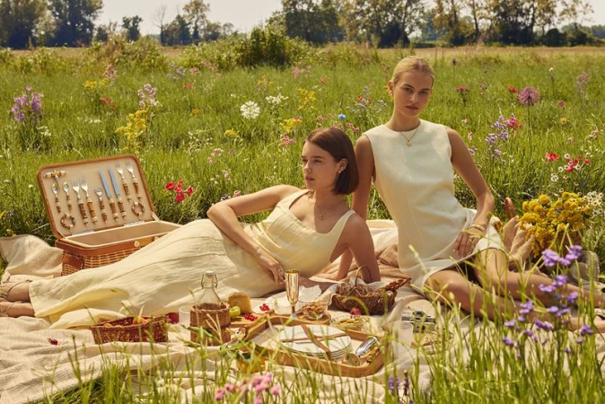 Two adult women enjoy a sunlit picnic in a wildflower meadow. They are seated on a blanket with a picnic basket and food.