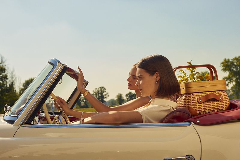 Two women drive a vintage convertible on a sunny day. The car is filled with a picnic basket and blanket, suggesting a road trip.