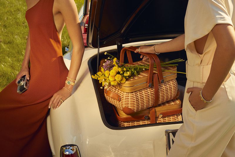 Two women load picnic baskets into a vintage car trunk in a sunlit field, wearing dresses and bracelets with an old camera visible.