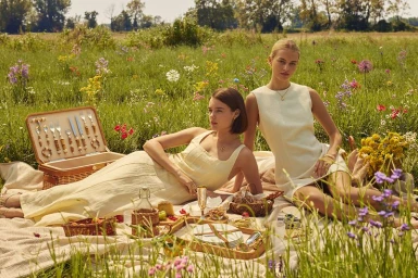 Two women in light cream dresses reclining on a picnic blanket in a sunlit wildflower field beside a wicker picnic hamper.
