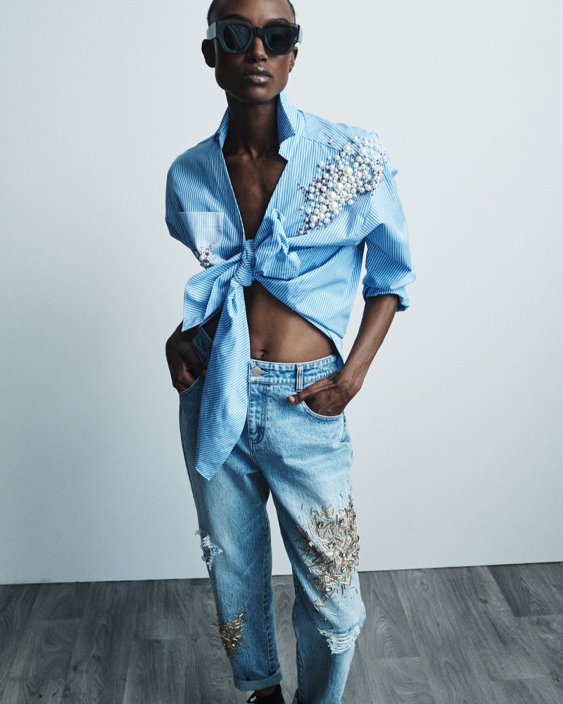 A woman in sunglasses wears a tied striped shirt and distressed jeans, accessorized with pearls and metallic embellishments, in a studio shot.