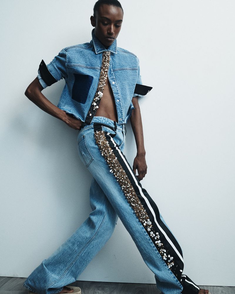 Fashionable, light-skinned person in a light denim outfit poses against a white wall. The outfit is embellished with gold and white details.