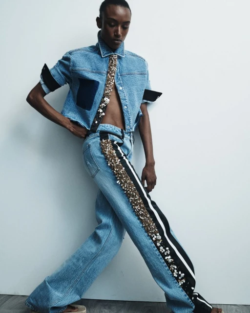 Fashionable, light-skinned person in a light denim outfit poses against a white wall. The outfit is embellished with gold and white details.