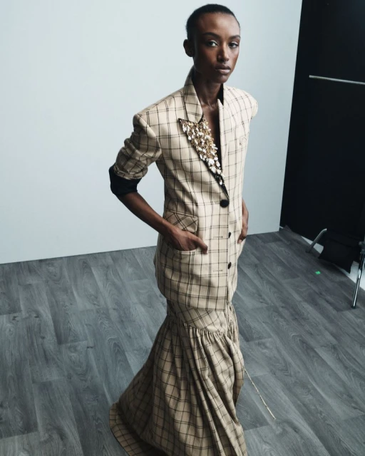 A black female model poses indoors wearing a tan and black plaid dress with a jacket-style top and a trumpet-style skirt and neutral background.