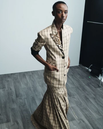 A black female model poses indoors wearing a tan and black plaid dress with a jacket-style top and a trumpet-style skirt and neutral background.