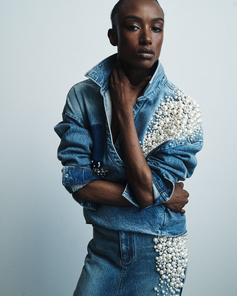 Portrait of a Black woman in a pearl-embellished denim jacket and skirt, striking a pose in a studio setting with soft, diffused lighting.