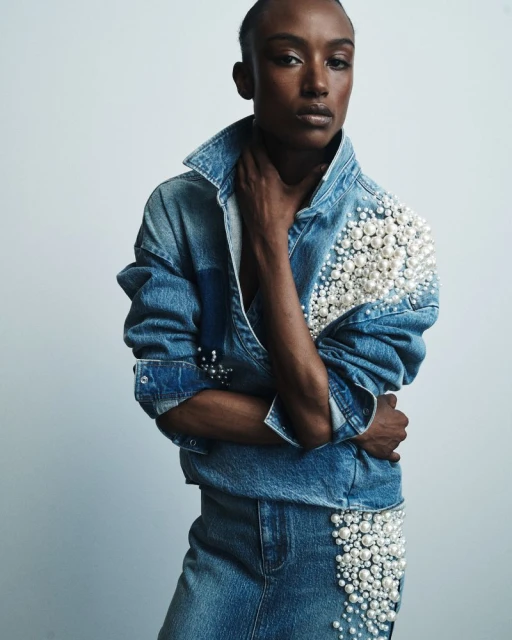 Portrait of a Black woman in a pearl-embellished denim jacket and skirt, striking a pose in a studio setting with soft, diffused lighting.