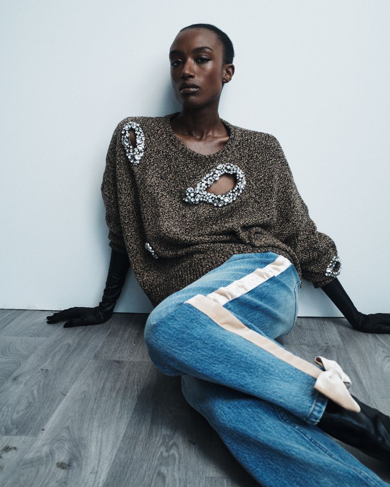 Dark-skinned woman wearing a brown sweater with cutouts, jeans with a beige stripe, and pointy-toe shoes in a fashion shot.