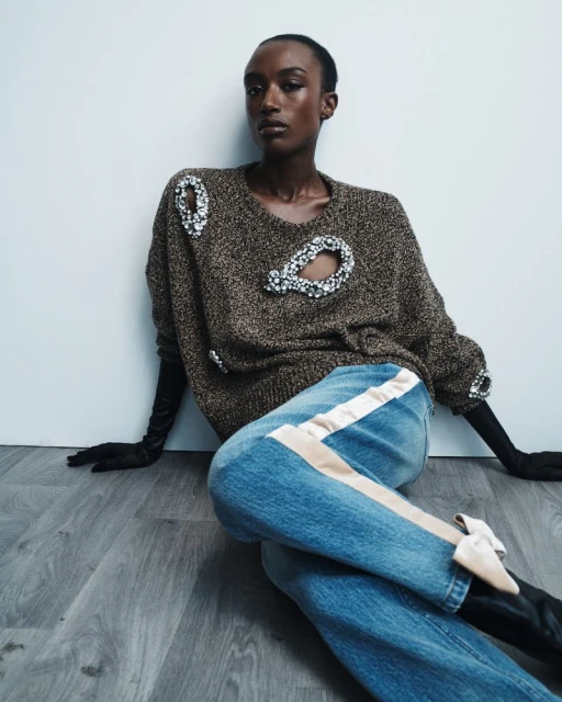 Dark-skinned woman wearing a brown sweater with cutouts, jeans with a beige stripe, and pointy-toe shoes in a fashion shot.