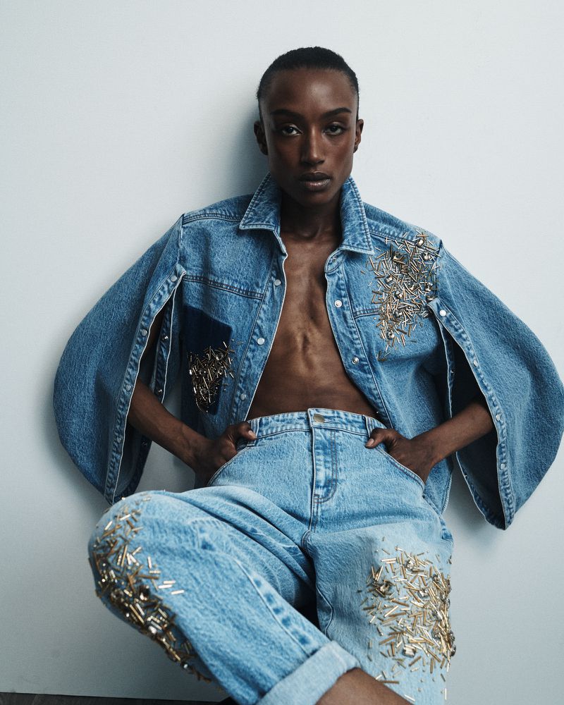 Elegant, dark-skinned model poses indoors in an embellished denim jacket and jeans with hands on waist against a white backdrop.