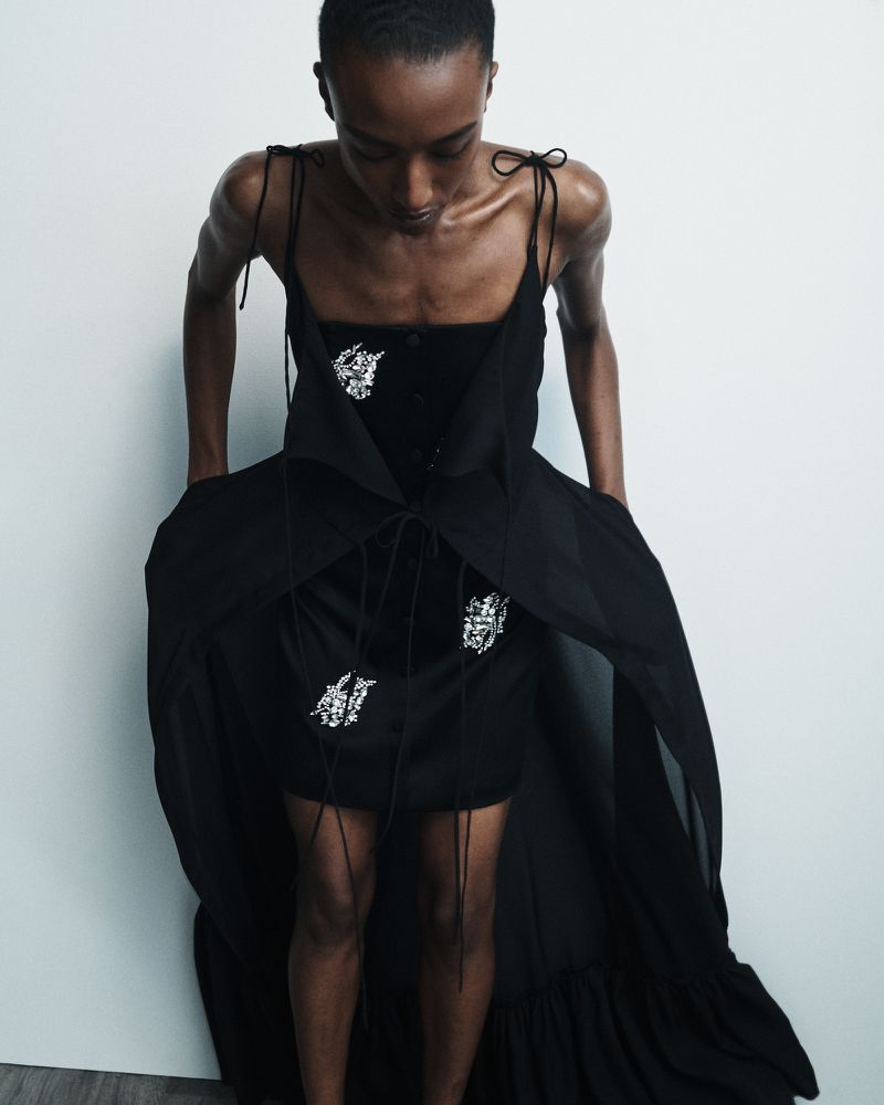 Fashion portrait of an adult woman in a black dress with thin straps, with a long black train, posing in front of a white wall.