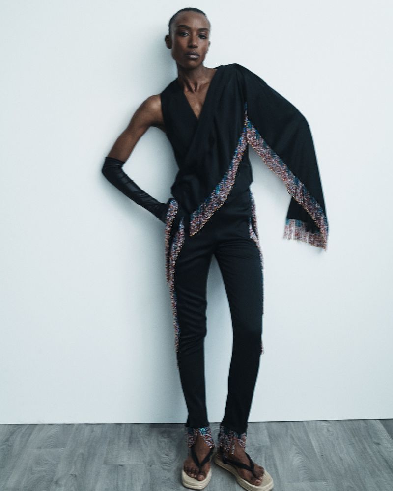 A slender fashion model in a striking black outfit, accessorized with beaded accents, poses against a white wall, showcasing unique style and design.
