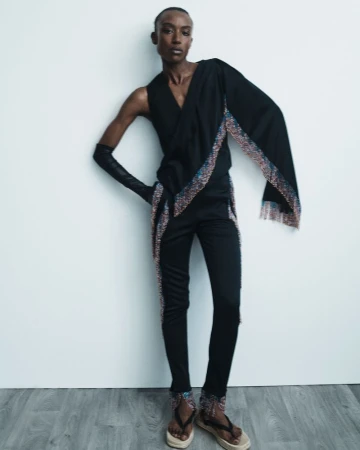 A slender fashion model in a striking black outfit, accessorized with beaded accents, poses against a white wall, showcasing unique style and design.