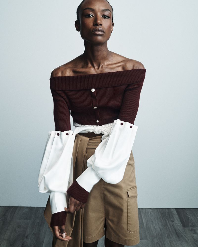 Portrait of a Black woman in a fashionable burgundy off-shoulder top with white billowy sleeves and khaki shorts in a studio setting.