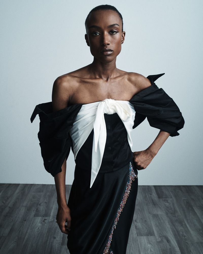 Elegant portrait of a Black woman in a black and white dress with a bow, posed in front of a light gray wall and wood floor.