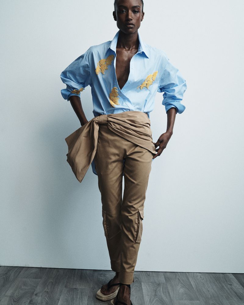 Fashion portrait of a Black woman in a blue striped shirt with gold embellishments and tan pants with a tie, posing indoors.