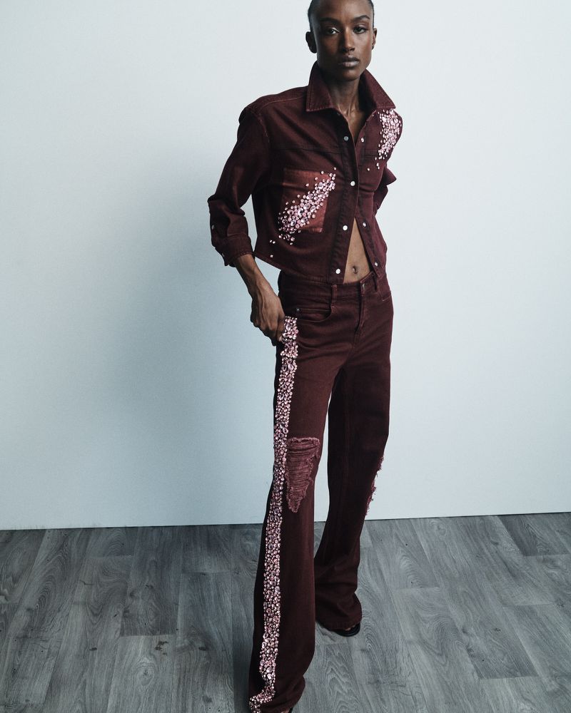 An adult woman poses in a studio. She wears a red denim jacket and matching jeans with a stripe. The floor is dark wood.