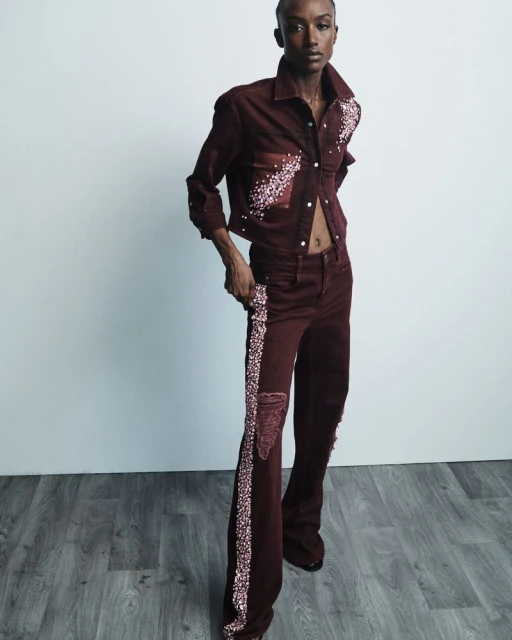 An adult woman poses in a studio. She wears a red denim jacket and matching jeans with a stripe. The floor is dark wood.