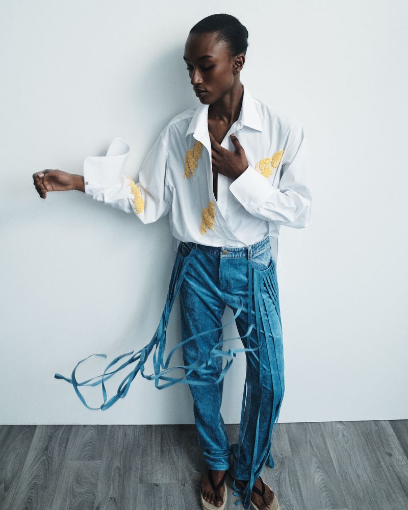 Stylish person in a white embroidered shirt and blue jeans with tassels posing in front of a white wall, showcasing fashion.