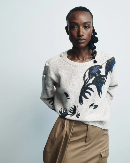 Studio shot of an African American woman wearing a cream colored sweater with a dark design and a brown skirt, posing against a white backdrop.