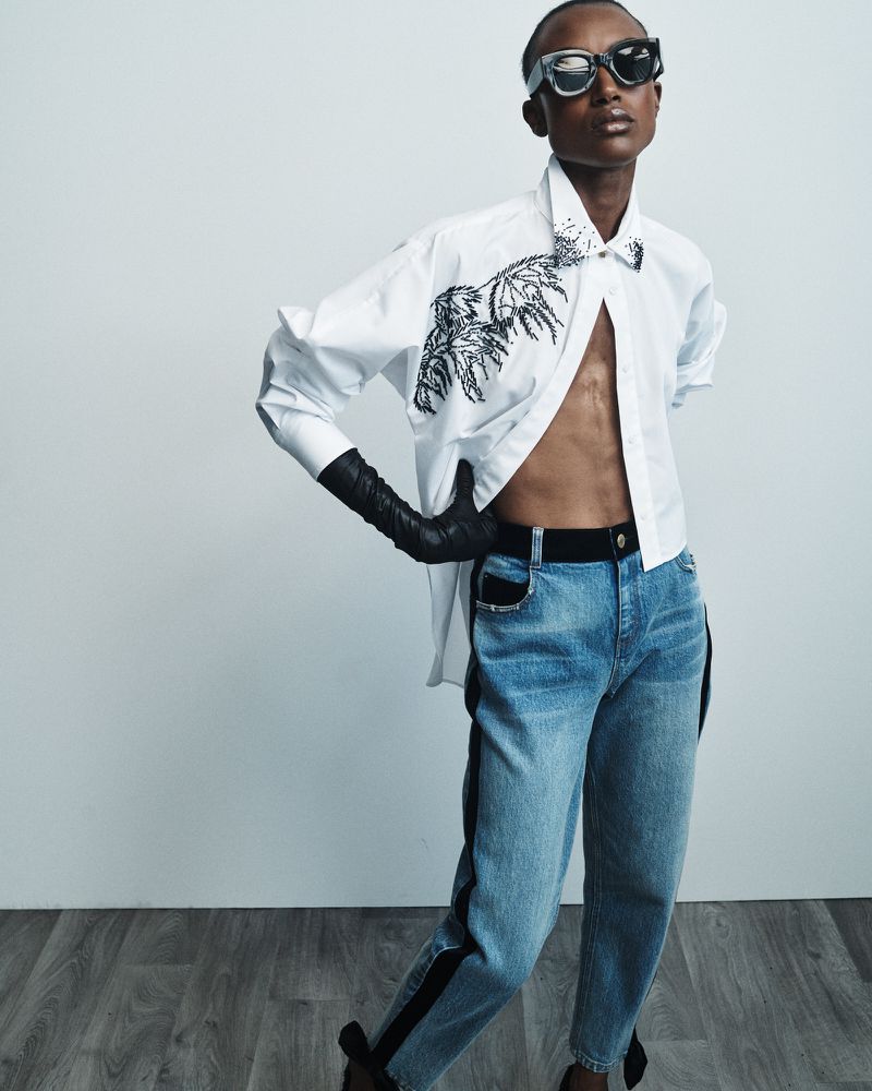 Stylish person poses in an unbuttoned shirt with an animal graphic, blue jeans with black stripe, sunglasses, and long black gloves.