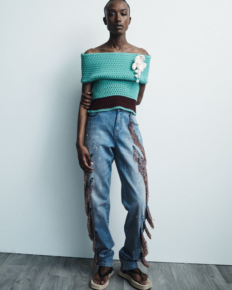 A woman poses in a teal off-the-shoulder top and fringed jeans against a white wall and gray floor in a fashion editorial studio shot.