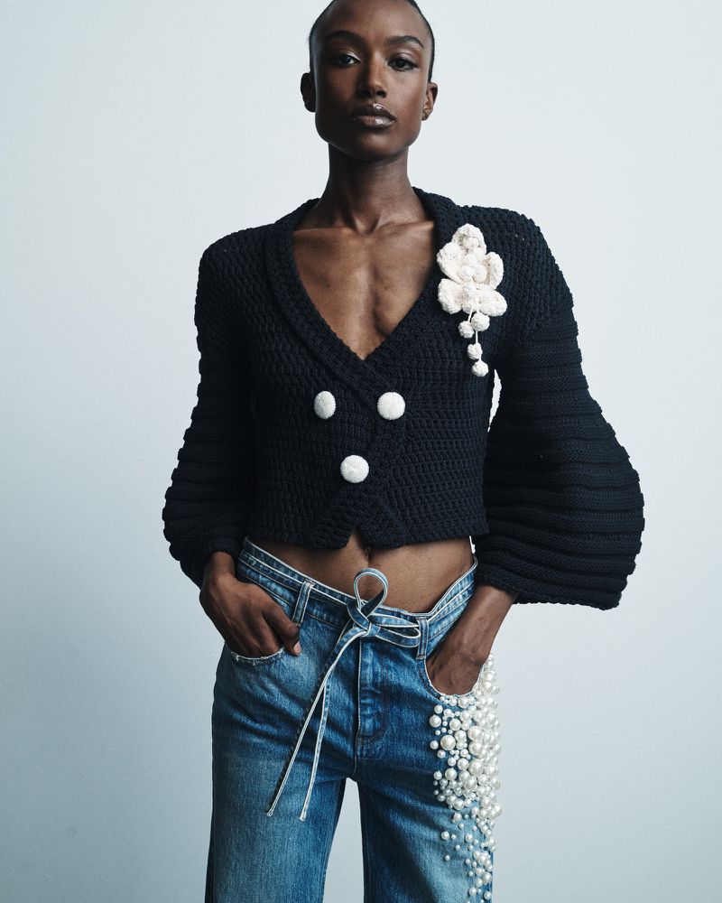 Adult female fashion model in black knit cardigan and denim jeans with pearl embellishments posing in a light grey studio setting.