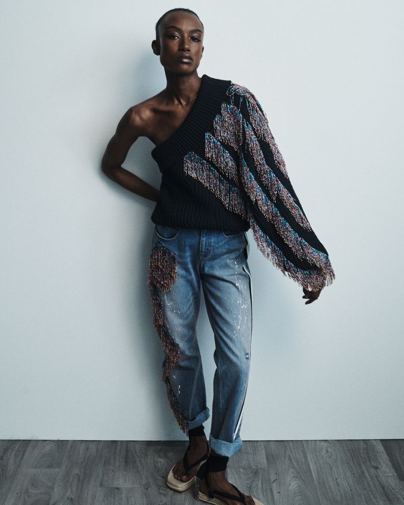 A Black female model poses in a studio wearing a black one-shoulder sweater with fringe, light wash jeans, and heels, leaning against a wall.