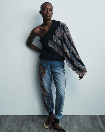 A Black female model poses in a studio wearing a black one-shoulder sweater with fringe, light wash jeans, and heels, leaning against a wall.