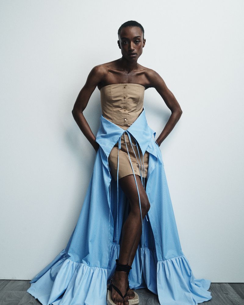 A Black fashion model poses in a tan tube top dress with a blue ruffled overlay against a white backdrop in a studio setting.