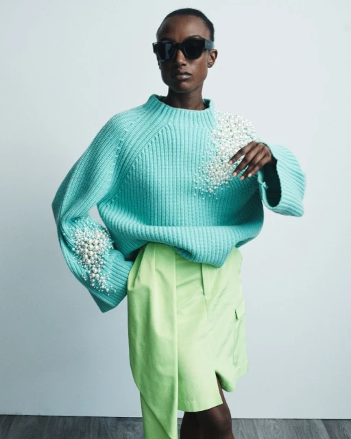 Fashion shot of a Black woman wearing a light blue knit sweater with pearl embellishments and bright green shorts against a white backdrop.