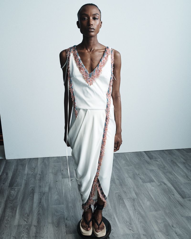 Full-length shot of a Black female fashion model wearing a white dress with colorful beaded trim, standing on a small platform.