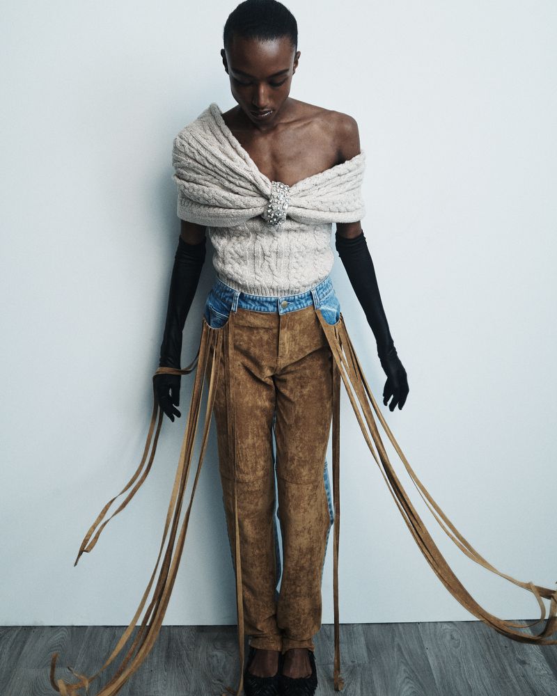 A slender African-American model poses in a creamy knit top, distressed blue jeans, and brown pants with fringe against a white background.