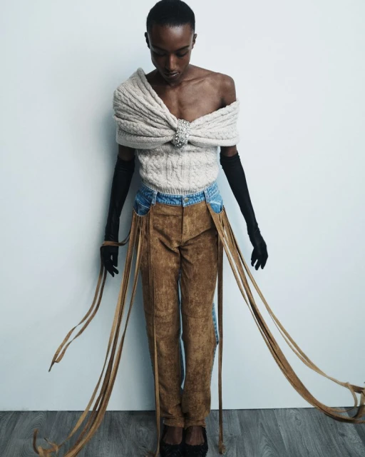 A slender African-American model poses in a creamy knit top, distressed blue jeans, and brown pants with fringe against a white background.