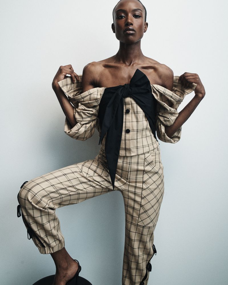 Fashion portrait of an African-descent adult female in plaid pants and a strapless top with bow detail, posing against a white background.