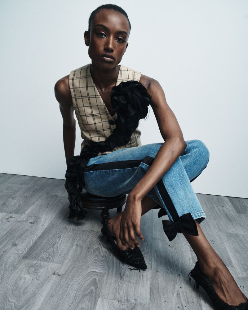 An adult woman in a plaid vest and jeans adorned with black bows is seated in a minimalist studio, creating a stylish, fashionable image.