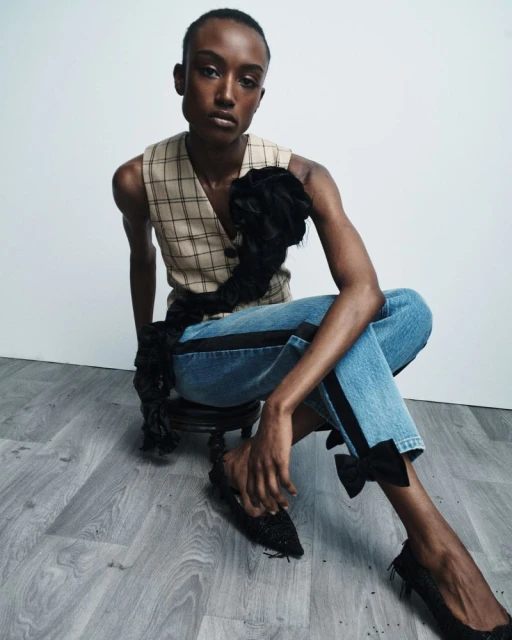 An adult woman in a plaid vest and jeans adorned with black bows is seated in a minimalist studio, creating a stylish, fashionable image.