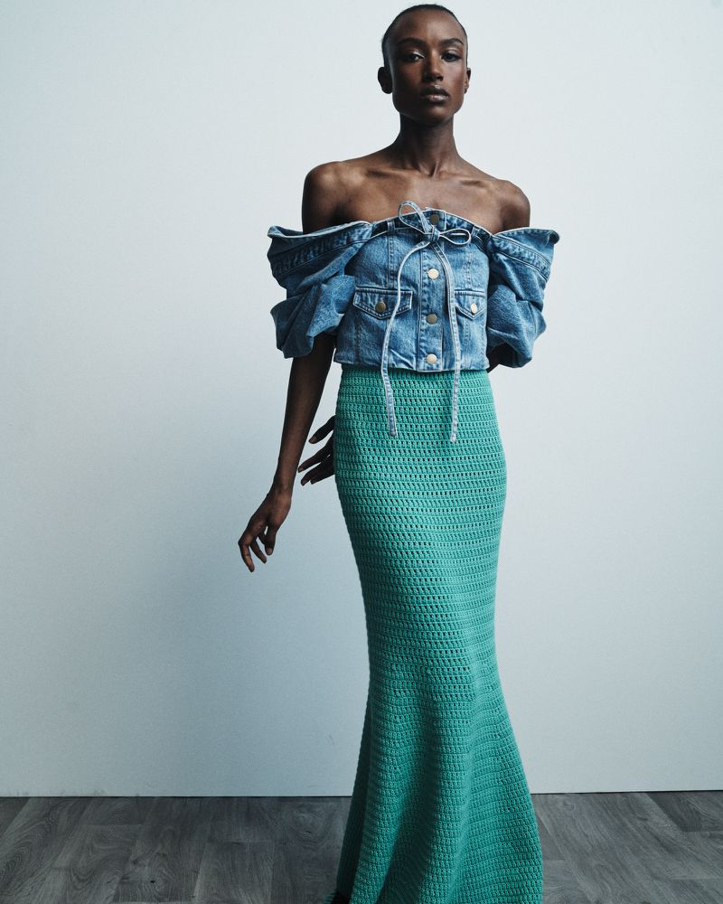 Dark-skinned model poses in off-the-shoulder denim top and crocheted turquoise skirt against a neutral backdrop in a fashion shot.