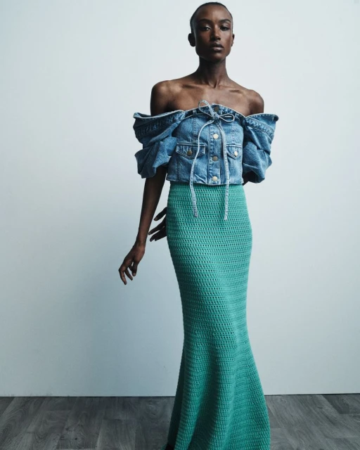 Dark-skinned model poses in off-the-shoulder denim top and crocheted turquoise skirt against a neutral backdrop in a fashion shot.