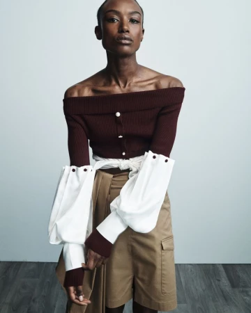 Model Amelie Nsengiyumva wearing a burgundy off-the-shoulder knit top with white accent sleeves and khaki cargo shorts.