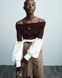 Model Amelie Nsengiyumva wearing a burgundy off-the-shoulder knit top with white accent sleeves and khaki cargo shorts.