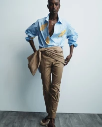 Model wearing a light blue button-up shirt and tan trousers standing against a pale studio wall